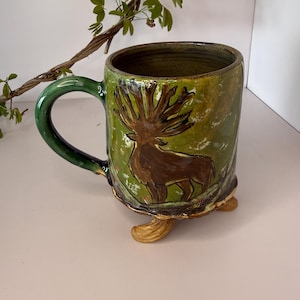 May include: Handmade ceramic mug with a green handle and a brown deer illustration on a green and yellow background. The mug has a unique design with three small feet and is perfect for coffee or tea.