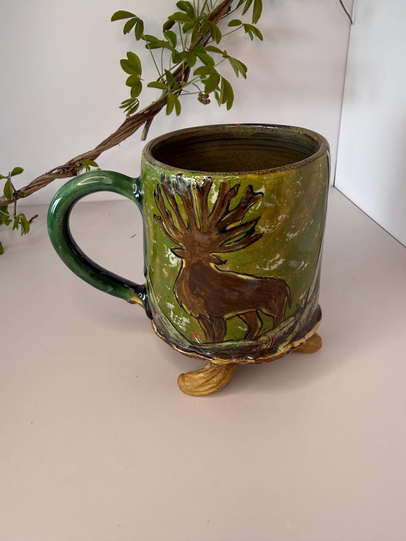 May include: Handmade ceramic mug with a green handle and a brown deer illustration on a green and yellow background. The mug has a unique design with three small feet and is perfect for coffee or tea.