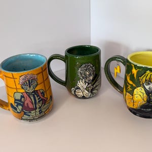 Puede incluir: Tres tazas de cer&aacute;mica con ilustraciones de personajes. Una taza es naranja con interior azul y un dise&ntilde;o de personaje. Otra es verde con un dise&ntilde;o de personaje. La tercera taza es verde con interior amarillo y un dise&ntilde;o de personaje, con un dije de rayo.