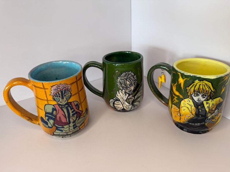 Puede incluir: Tres tazas de cer&aacute;mica con ilustraciones de personajes. Una taza es naranja con interior azul y un dise&ntilde;o de personaje. Otra es verde con un dise&ntilde;o de personaje. La tercera taza es verde con interior amarillo y un dise&ntilde;o de personaje, con un dije de rayo.