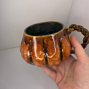 May include: A ceramic mug shaped like a pumpkin with a glossy orange and brown glaze. The mug features vertical ridges mimicking a pumpkin's texture and a dark interior. It includes a handle designed with a twisted, rope-like aesthetic.