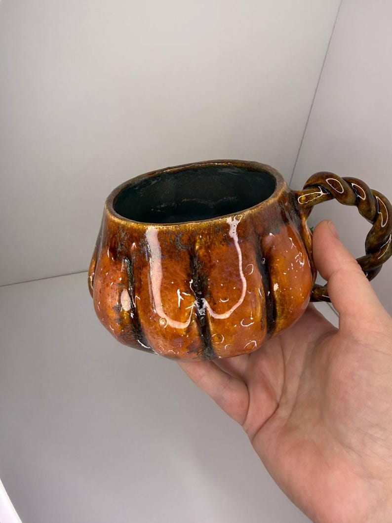 May include: A ceramic mug shaped like a pumpkin with a glossy orange and brown glaze. The mug features vertical ridges mimicking a pumpkin's texture and a dark interior. It includes a handle designed with a twisted, rope-like aesthetic.