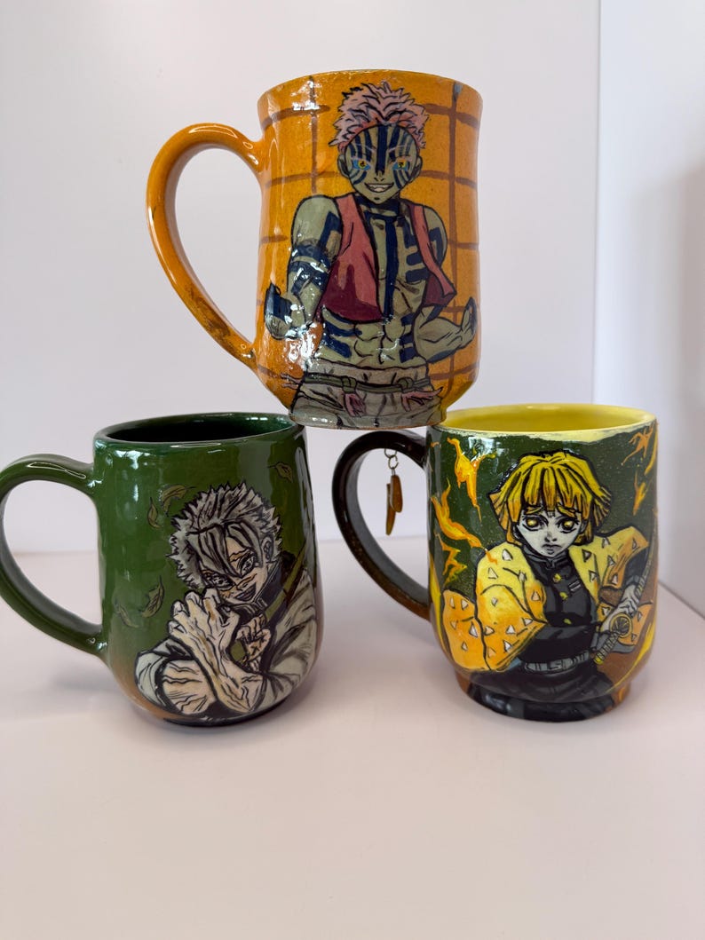 Puede incluir: Tres tazas de cer&aacute;mica con ilustraciones de personajes. La taza superior es naranja con un personaje con chaleco rojo. La taza del medio es verde con una ilustraci&oacute;n de personaje. La taza inferior es amarilla con una ilustraci&oacute;n de personaje y un asa negra.