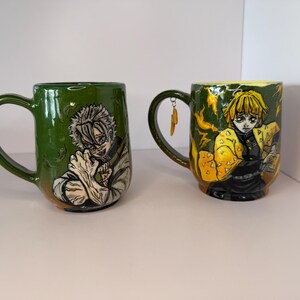 Puede incluir: Dos tazas de cer&aacute;mica con ilustraciones de personajes de anime. Una es verde con un personaje de pelo blanco, la otra es verde y amarilla con un personaje de pelo amarillo. Ambas tazas tienen asas grandes y est&aacute;n sobre una superficie blanca.
