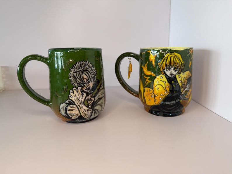 Puede incluir: Dos tazas de cer&aacute;mica con ilustraciones de personajes de anime. Una es verde con un personaje de pelo blanco, la otra es verde y amarilla con un personaje de pelo amarillo. Ambas tazas tienen asas grandes y est&aacute;n sobre una superficie blanca.