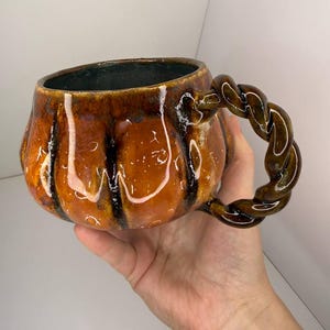 May include: A ceramic mug shaped like a pumpkin with a glossy orange and brown glaze. The mug features vertical ridges and a dark interior. It has a distinctive twisted, rope-like handle attached to the side, finished in a matching dark brown glaze.