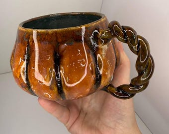 May include: A ceramic mug shaped like a pumpkin with a glossy orange and brown glaze. The mug features vertical ridges and a dark interior. It has a distinctive twisted, rope-like handle attached to the side, finished in a matching dark brown glaze.