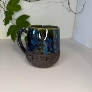 May include: A ceramic mug featuring a glossy, mottled blue and green glaze on the upper portion and a dark, matte brown base. The lower section is embossed with a band of runic characters. The mug has a curved handle and a rounded body shape.