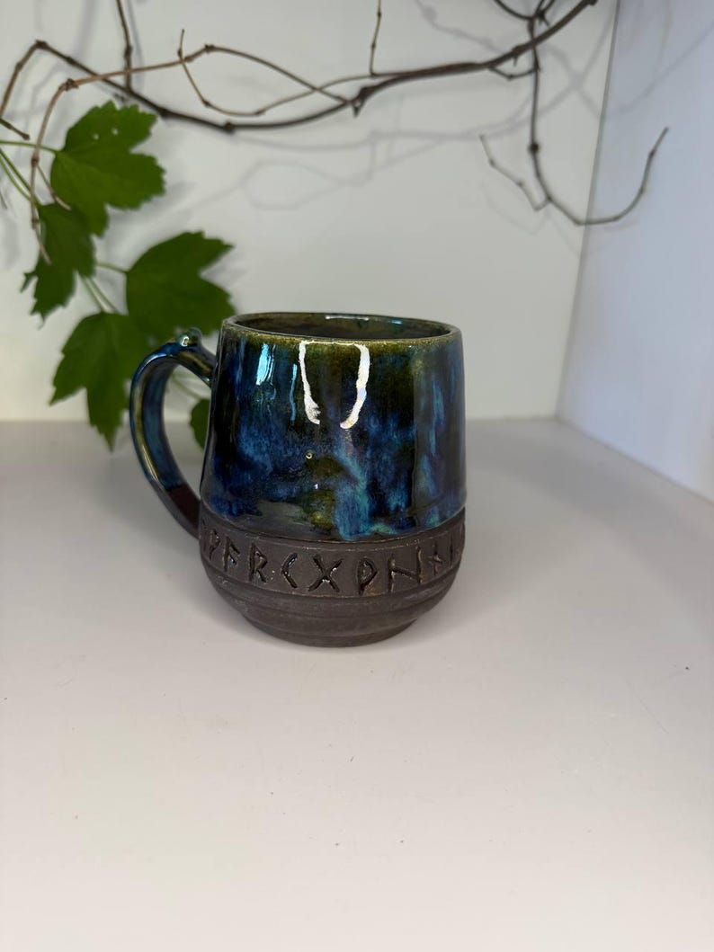 May include: A ceramic mug featuring a glossy, mottled blue and green glaze on the upper portion and a dark, matte brown base. The lower section is embossed with a band of runic characters. The mug has a curved handle and a rounded body shape.