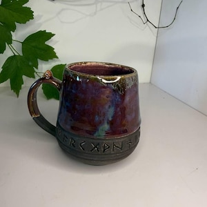 May include: A ceramic mug with a glossy, multi-colored glaze in shades of deep purple, blue, and brown. The lower portion of the mug features a matte black band embossed with a series of runic characters. It has a sturdy handle and a wide rim.