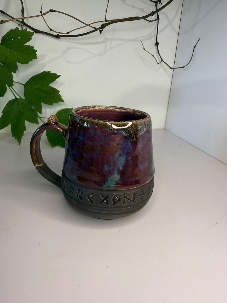 May include: A ceramic mug with a glossy, multi-colored glaze in shades of deep purple, blue, and brown. The lower portion of the mug features a matte black band embossed with a series of runic characters. It has a sturdy handle and a wide rim.