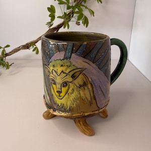 May include: A ceramic mug with a whimsical design. The mug features a hand-painted illustration of a yellow creature with purple accents and a green handle. The mug is supported by three small, tan-colored feet.