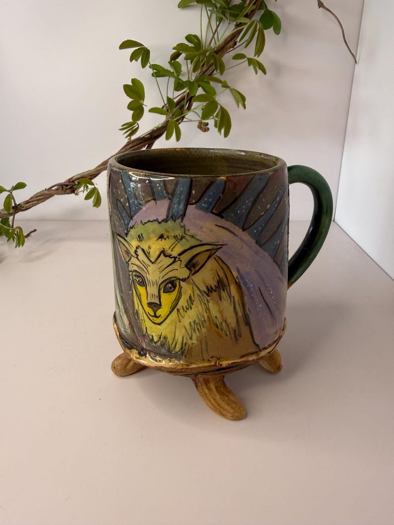 May include: A ceramic mug with a whimsical design. The mug features a hand-painted illustration of a yellow creature with purple accents and a green handle. The mug is supported by three small, tan-colored feet.