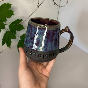 May include: A ceramic mug with a dark, matte black base featuring the embossed text "MYSTMPA" in a runic-style font. The upper portion of the mug has a glossy, mottled glaze in shades of deep purple, blue, and green, with a curved handle.