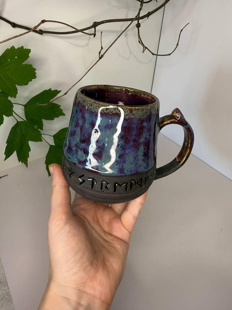 May include: A ceramic mug with a dark, matte black base featuring the embossed text "MYSTMPA" in a runic-style font. The upper portion of the mug has a glossy, mottled glaze in shades of deep purple, blue, and green, with a curved handle.