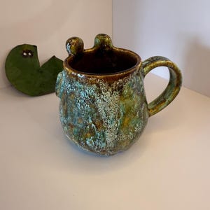 May include: A ceramic mug with a rounded body and a curved handle, featuring a mottled glaze in shades of deep brown, forest green, and turquoise. The rim has two small, rounded protrusions extending upward, giving the vessel a unique silhouette.