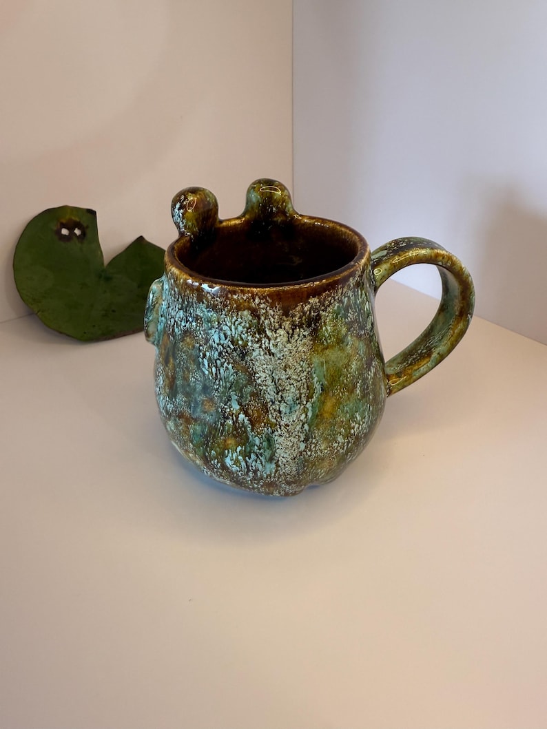 May include: A ceramic mug with a rounded body and a curved handle, featuring a mottled glaze in shades of deep brown, forest green, and turquoise. The rim has two small, rounded protrusions extending upward, giving the vessel a unique silhouette.