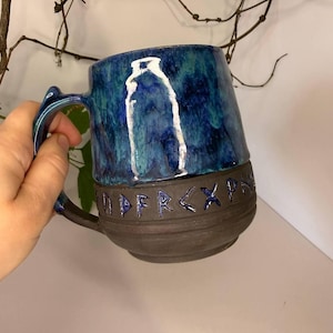 May include: A ceramic mug with a dark brown base and a glossy blue glazed upper section. The mug features a white outline of a bottle on the front and a band of carved runic characters around the middle. It has a curved handle on the side.
