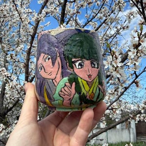 Könnte beinhalten: Ein Keramikbecher mit einer handgemalten Illustration von zwei Anime-Figuren. Eine Figur hat langes violettes Haar, die andere kurzes grünes Haar, Sommersprossen und einen gelben Kragen. Der Becher hat eine texturierte, gesprenkelte Oberfläche.