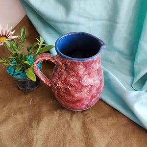 Peut inclure: Une cruche en céramique faite à la main avec un extérieur rouge et blanc moucheté et un intérieur bleu. La cruche a une anse et un bec verseur. Une petite plante en pot avec une fleur blanche et violette est à côté de la cruche.