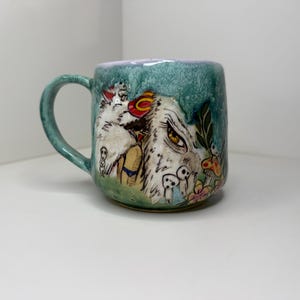May include: A ceramic mug with a teal and lavender glaze featuring a hand-painted illustration of a white, furry creature with yellow eyes, small forest spirits, mushrooms, and foliage. The mug has a curved handle and a rounded base.