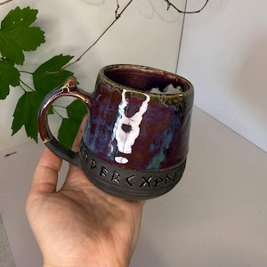 May include: A ceramic mug with a glossy, multi-colored glaze in shades of deep purple, blue, and green on the upper portion. The lower section is matte black and features a band of carved runic characters encircling the base of the vessel.