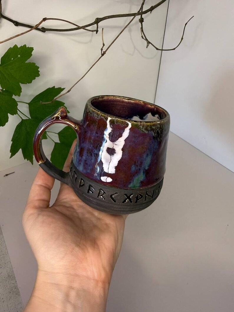 May include: A ceramic mug with a glossy, multi-colored glaze in shades of deep purple, blue, and green on the upper portion. The lower section is matte black and features a band of carved runic characters encircling the base of the vessel.