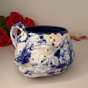 May include: A ceramic mug with a sculpted face featuring a blue and white marbled glaze. The surface is decorated with small, embedded gold-colored circular accents. The interior of the mug is a solid deep blue, and it includes a curved handle.