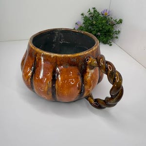 May include: A ceramic mug shaped like a pumpkin with a glossy orange and brown glaze. The mug features vertical ridges along its rounded body and a distinctive twisted, rope-like handle attached to the side. The interior is dark and smooth.