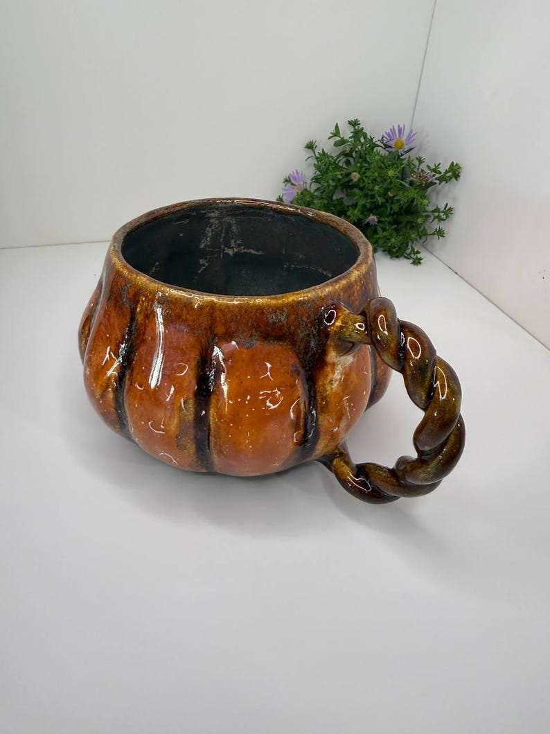 May include: A ceramic mug shaped like a pumpkin with a glossy orange and brown glaze. The mug features vertical ridges along its rounded body and a distinctive twisted, rope-like handle attached to the side. The interior is dark and smooth.