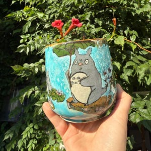 May include: A ceramic mug featuring a hand-painted illustration of a grey, round creature with a white belly holding a large green leaf as an umbrella. The background of the mug is a textured, bright turquoise blue with smaller grey figures.