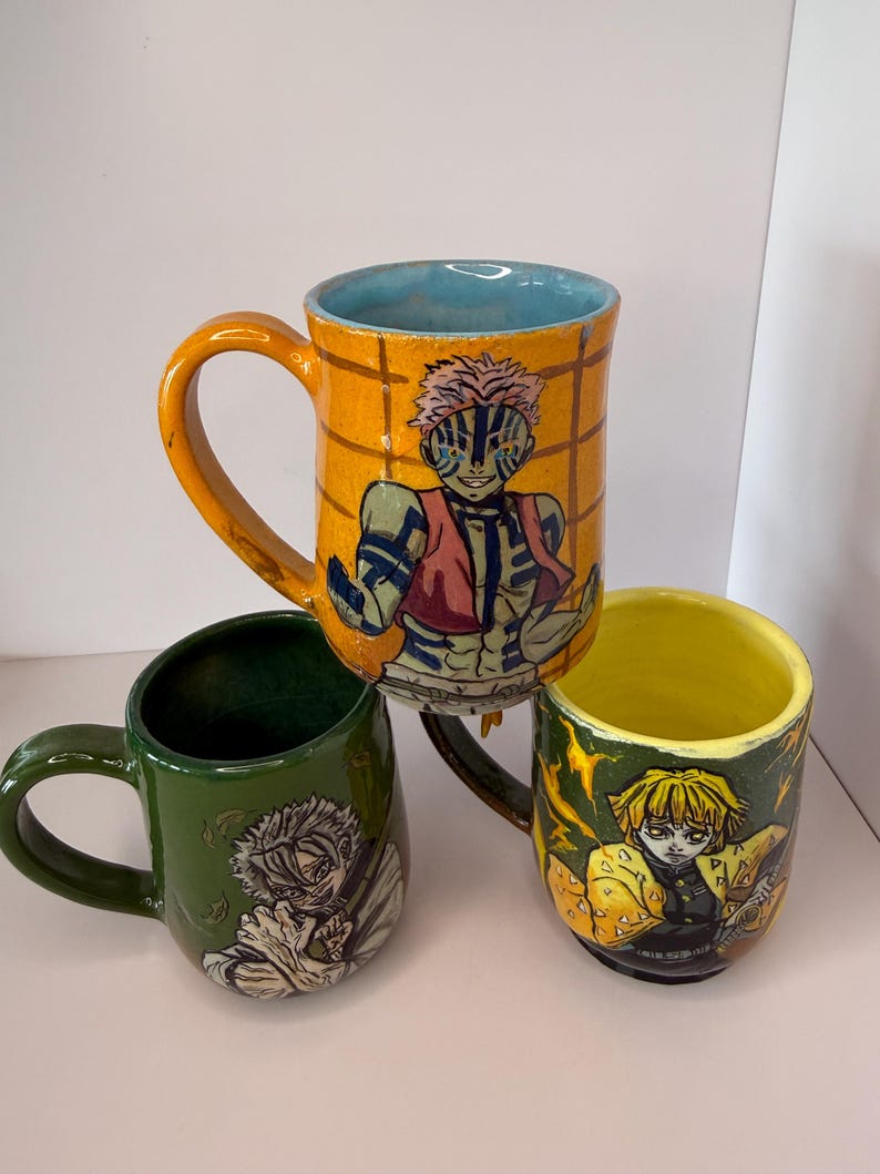 Puede incluir: Tres tazas de cer&aacute;mica con ilustraciones de personajes. Una taza es naranja con interior azul y dise&ntilde;o de personaje. Otra es verde con dise&ntilde;o de personaje. La tercera es amarilla con dise&ntilde;o de personaje.