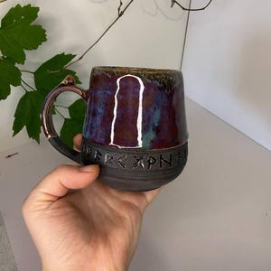 May include: A ceramic mug with a dark matte base and a glossy, multi-colored glaze in shades of purple, blue, and brown. The mug features a band of carved runic characters around the middle and a single handle with a small, pointed protrusion.