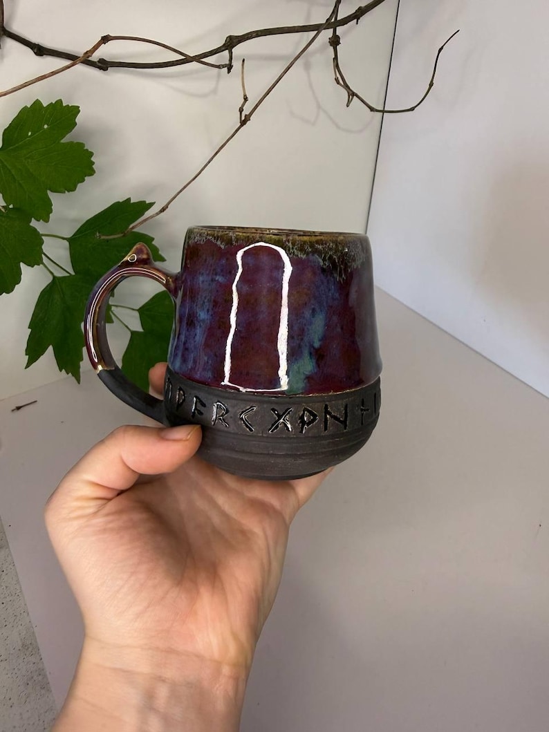 May include: A ceramic mug with a dark matte base and a glossy, multi-colored glaze in shades of purple, blue, and brown. The mug features a band of carved runic characters around the middle and a single handle with a small, pointed protrusion.