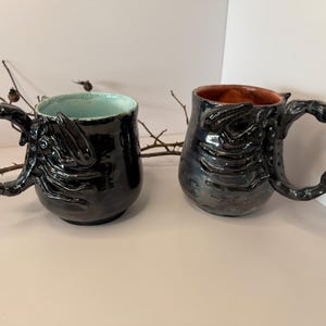 May include: Two handmade ceramic mugs. The mugs are black with textured designs and unique handles. One mug has a light blue interior, and the other has a brown interior. The mugs are displayed on a neutral surface.