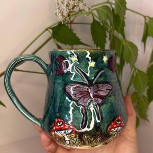 Może przedstawiać: Ręcznie wykonany ceramiczny kubek z błyszczącym, turkusowo-zielonym szkliwem. Powierzchnia ozdobiona jest wypukłymi wzorami, w tym centralnie umieszczoną fioletową ćmą, dwoma czerwono-białymi grzybami u podstawy oraz małymi żółtymi gwiazdkami i księżycami.
