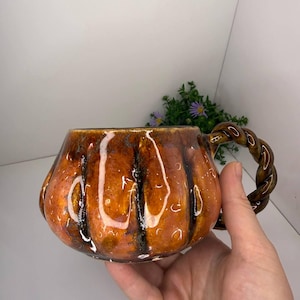 May include: A ceramic mug shaped like a pumpkin with a glossy orange and brown glaze. The mug features vertical ridges mimicking a pumpkin's texture and a dark brown, twisted rope-style handle attached to the side of the rounded vessel.
