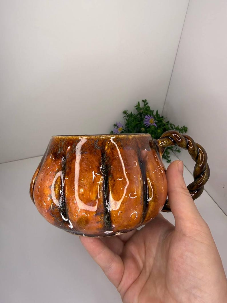May include: A ceramic mug shaped like a pumpkin with a glossy orange and brown glaze. The mug features vertical ridges mimicking a pumpkin's texture and a dark brown, twisted rope-style handle attached to the side of the rounded vessel.