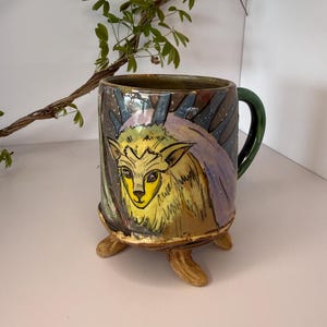 May include: Hand-painted ceramic mug with a whimsical deer design. The mug features a yellow deer face with a green handle and three brown feet. The background has a dark, starry night design with purple and blue accents. A unique, artistic piece.