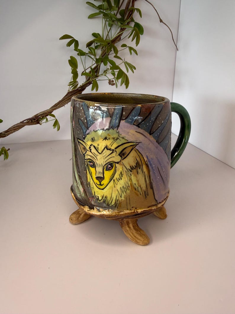 May include: Hand-painted ceramic mug with a whimsical deer design. The mug features a yellow deer face with a green handle and three brown feet. The background has a dark, starry night design with purple and blue accents. A unique, artistic piece.