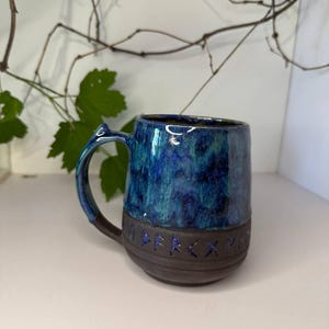 May include: A ceramic mug featuring a glossy, mottled blue and green glaze on the upper portion and a matte dark brown base. The lower section is embossed with a series of runic characters highlighted in blue. It includes a curved handle.