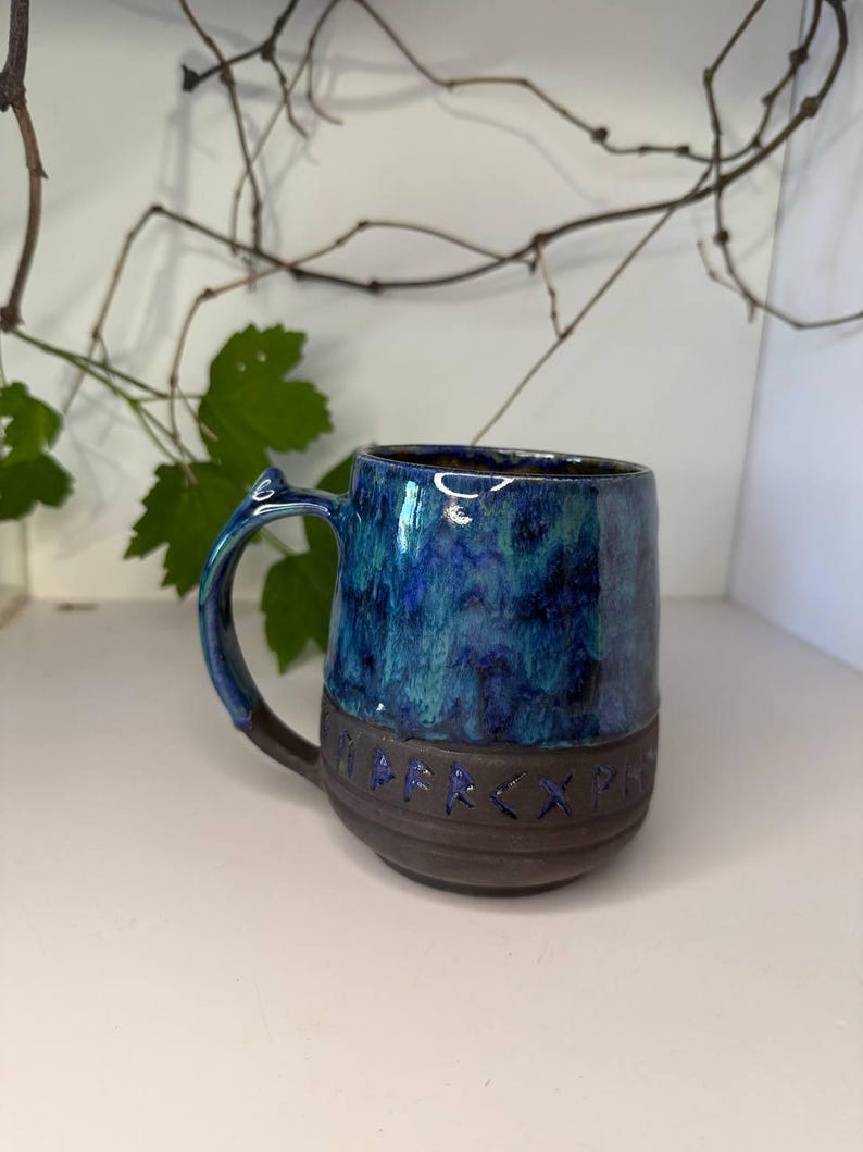 May include: A ceramic mug featuring a glossy, mottled blue and green glaze on the upper portion and a matte dark brown base. The lower section is embossed with a series of runic characters highlighted in blue. It includes a curved handle.