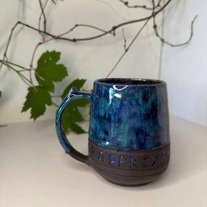 May include: A ceramic mug featuring a glossy, mottled blue and teal glaze on the upper portion and a matte dark brown base. The lower section is embossed with a series of runic characters. The mug has a curved handle with a small pointed protrusion.