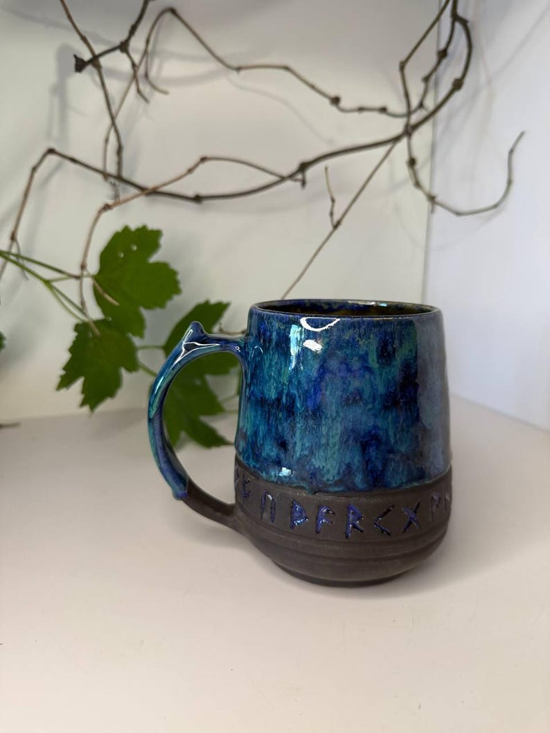 May include: A ceramic mug featuring a glossy, mottled blue and teal glaze on the upper portion and a matte dark brown base. The lower section is embossed with a series of runic characters. The mug has a curved handle with a small pointed protrusion.