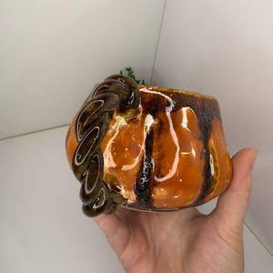 May include: A ceramic mug with a glossy orange glaze and dark brown vertical drip accents. The mug features a distinctive handle shaped like a thick, twisted rope in a dark brown finish, providing a textured contrast to the smooth, rounded body.