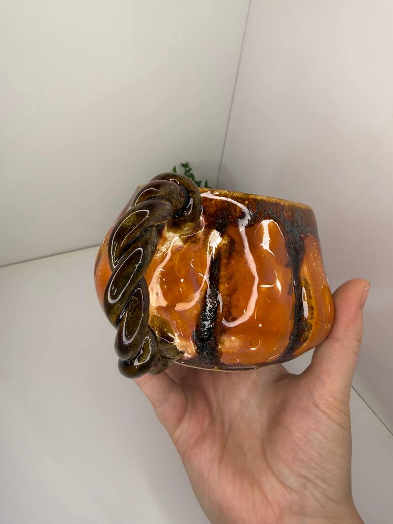 May include: A ceramic mug with a glossy orange glaze and dark brown vertical drip accents. The mug features a distinctive handle shaped like a thick, twisted rope in a dark brown finish, providing a textured contrast to the smooth, rounded body.