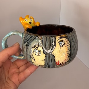 Könnte beinhalten: Eine Keramik-Tasse mit hellblauem Henkel und detailliertem Design. Die Tasse zeigt zwei Gesichter und eine kleine, gelbe, cartoonartige Figur, die auf dem Rand sitzt. Das Innere der Tasse ist dunkelbraun.