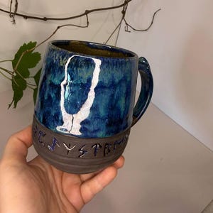 May include: A ceramic mug featuring a glossy blue and teal glaze on the upper portion and a matte dark brown base. The mug displays a white abstract silhouette graphic and embossed lettering that reads "PLAYSYSTEM" around the lower section.
