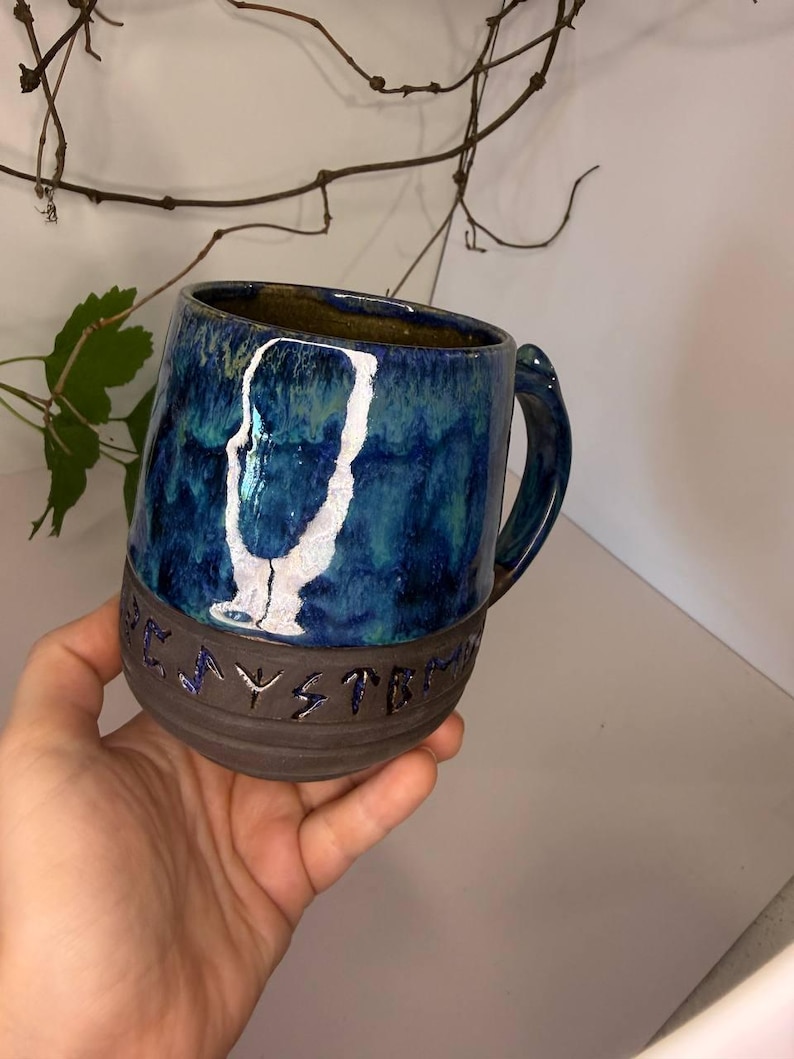 May include: A ceramic mug featuring a glossy blue and teal glaze on the upper portion and a matte dark brown base. The mug displays a white abstract silhouette graphic and embossed lettering that reads "PLAYSYSTEM" around the lower section.