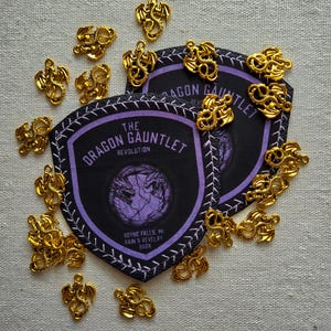 May include: Two shield-shaped black and purple patches with the text THE DRAGON GAUNTLET REVOLUTION, BOYNE FALLS, MI, RAIN & REVELRY 2026, surrounded by numerous small, gold-colored metal dragon charms with intricate, coiled wing and tail details.